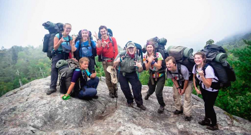 A group of people wearing backpacks stand on rocky ground, high above trees